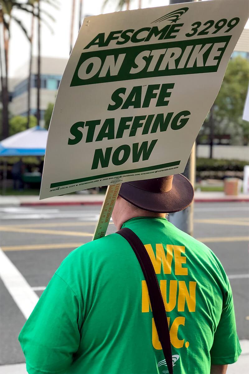 UC worker with back to the camera wearing a green shirt that reads, "We Run UC" and a sign, "AFSCME 3299 On Strike. Safe Staffing Now."
